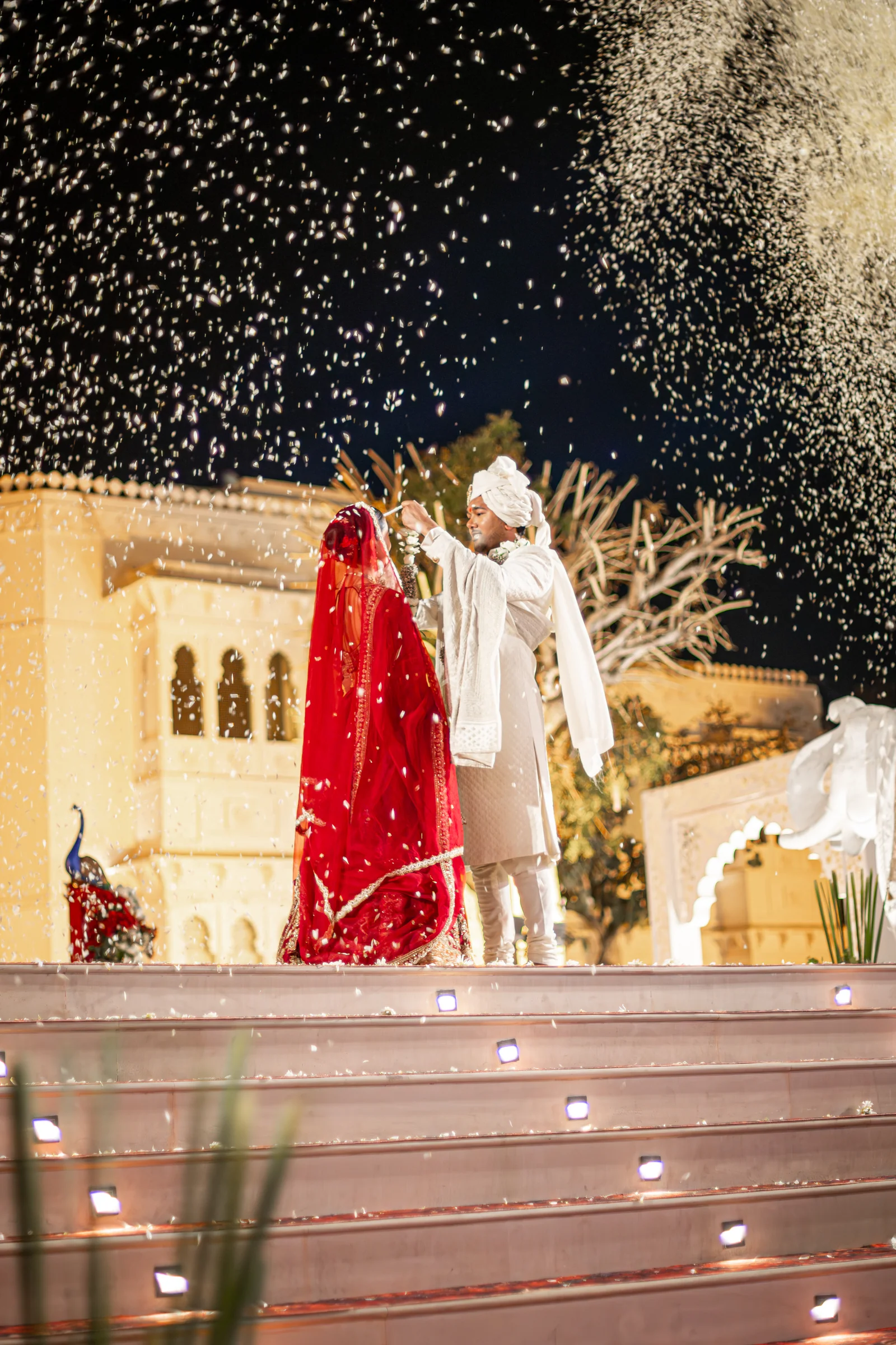 Jay and Serene stand beneath a shower of white petals at night